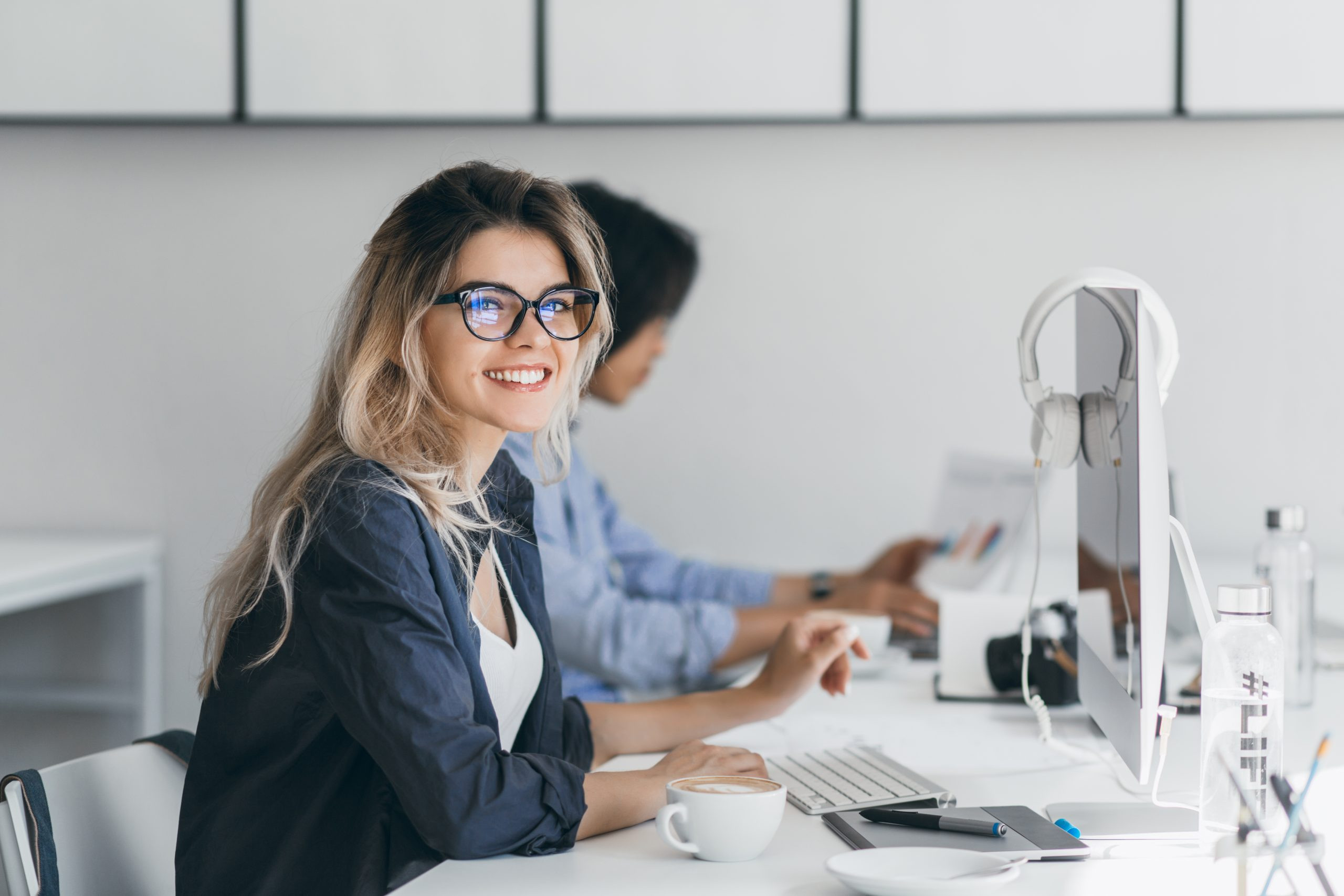 Attractive laughing freelancer girl posing with cup of coffee at her workplace. Chinese student in blue shirt works with document in campus with blonde friend in glasses..